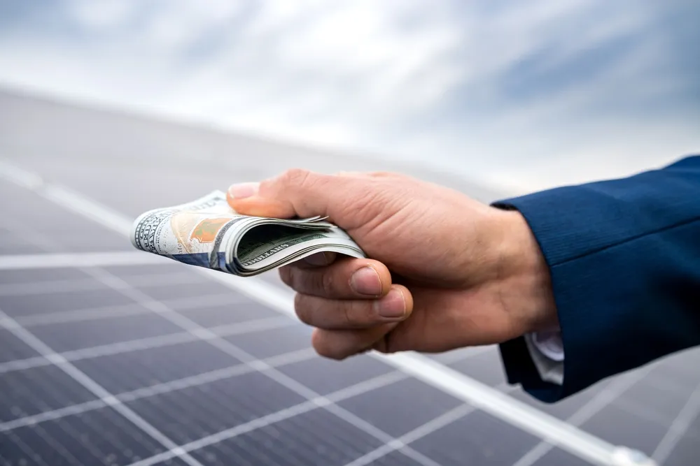 A businessman's hand holding money against solar panel installation.